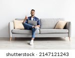 I Like. Portrait Of Happy Excited Middle Eastern Man Sitting On Couch Using Laptop And Showing Thumbs Up Sign Gesture, Working Or Studying Remotely, Posing To Camera In Living Room, Full Body Length
