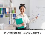 Small photo of Training concept. Happy female tutor teaching English language, pointing at grammar rules on board and smiling at camera, holding clipboard in office interior