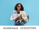 Online education. Smiling black female student with backpack and headphones holding notebooks and digital tablet on blue studio background. Elearning, studying remotely concept
