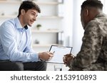 Small photo of Young man in formal outwear psychologist giving african american military man recommendations, showing list, soldier and psychotherapist having conversation, clinic interior