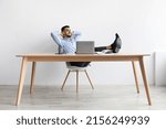 Relief After Finished Work. Smiling businessman relaxing on chair sitting at table resting feet on table, using pc, happy male leaning back at workplace holding hands behind head, feeling pleased