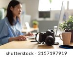 Asian Photographer Lady Using Laptop Sitting At Workplace, Typing On Keyboard, Selective Focus On Professional Photo Camera Lying On Table. Female Artist Transferring Pictures To Computer For Retouch