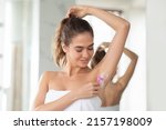 Young Lady Shaving Armpits Covered With Foam Removing Hair Standing Wrapped In Towel In Modern Bathroom. Woman Raising Arm Making Underarms Depilation. Hair Removal Concept