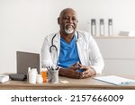 Small photo of Healthcare And Medicine Concept. Portrait of cheerful mature African American doctor posing, smiling looking at camera, sitting at desk in office. Trustworthy specialist in stethoscope and white coat