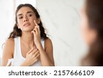 Small photo of Skin Problem. Depressed Woman Touching Pimple On Face Looking At Mirror In Modern Bathroom. Facial Skin Issues, Medical Care And Treatment Concept. Selective Focus