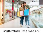 Small photo of Mobile Shopping. Couple Of Buyers Using Phone And Credit Card Purchasing New Clothes In Application Holding Colorful Shopper Bags Standing In Modern Mall. Ecommerce Offers And Sales