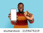 Online Trading. Excited Black Man Showing Golden Bitcoin Coin And Blank Smartphone With White Screen At Camera, African American Guy Advertising Crypto App While Posing On Blue Background, Mockup