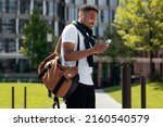 Stylish Young Black Man In Wireless Headphones Using Cell Phone Walking Down The Street In The Urban City. Cheerful Trendy Guy Listening To Favorite Music Spending Time Outdoors, Walking Resting Alone
