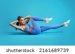 Small photo of Fitness Workout. Sportswoman In Fitwear Doing Elbow To Knee Abs Crunch Exercising On Blue Studio Background. Athletic Female Flexing Abs Muscles Lying On Floor. Sport And Bodybuilding