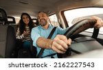 Small photo of Portrait of happy male driver riding car, cheerful beautiful lady sitting inside auto on back passenger seat, looking on the road abd talking with guy, enjoying safe ride, windshield view