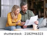 Small photo of Married Middle Aged Couple Planning Budget Together, Reading Papers And Calculating Spends While Sitting On Couch In Living Room, Husband And Wife Checking Documents And Accounting Taxes, Closeup