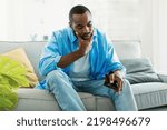Small photo of I'm broke. Sad african american man bankrupt showing his empty wallet and looking at it, sitting on sofa at home, free space. Poverty concept