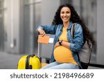 Small photo of Giving birth in foreign country concept. Happy young long-haired brunette pregnant woman travelling alone, sitting on bench with backpack next to baggage, holding passport, flight tickets, copy space