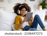 Small photo of Happy african american kid school girl with bushy hair sitting on couch, using digital tablet and wireless headset, playing mobile game or checking newest app, home interior, copy space