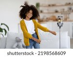 Small photo of Cute african american preteen girl with bushy hair in casual outfit recording video while dancing, showing dance moves, using cell phone on tripod, home interior, copy space. Kids lifestyle concept