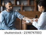 Small photo of Middle eastern young man having regular checkup at general practitioner at modern clinic, african american woman doctor checking heart rate for male patient suffering from hypertension, copy space