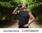 Small photo of Sporty young black man drinking protein cocktail, training at public park, using wireless earbuds, listening to music while jogging by forest, copy space. Sports nutrition concept