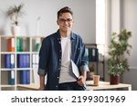 Portrait Of Handsome Young Male Entrepreneur Holding Laptop And Posing In Office Interior, Confident Millennial Man In Casual Clothes Standing Near Desk And Smiling At Camera, Copy Space
