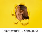 Small photo of Black man's ear and hand through a torn hole in yellow paper background, free copy space, closeup. The concept of eavesdropping, espionage, gossip and tabloids