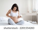 Small photo of Positive young pregnant woman in comfortable homewear sitting on bed, applying belly butter on her big tummy, white heart shaped symbol on pregnant woman belly, panorama with copy space, home interior