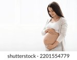 Small photo of Happy young pregnant woman in comfortable homewear standing next to window at home, looking at her big tummy, waiting for baby, panorama with copy space. Pregnancy, motherhood concept