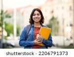 Small photo of Beautiful Young Arab Female Student Standing Outdoors With Workbooks In Hands, Happy Attractive Middle Eastern Woman Posing Outside On City Street, Carrying Backpack And Smiling At Camera, Copy Space
