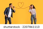 Small photo of Message With Love. Happy Young Arab Man Telling Romantic Words To His Girlfriend Through Tin Phone With Drawn Red Heart As String, Loving Middle Eastern Couple Posing Over Yellow Background