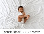 Cute Funny Little Black Baby Having Fun While Lying On Bed At Home, Adorable African American Infant Boy Playing With His Toes And Laughing While Resting In Bedroom, Top View, Copy Space