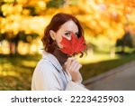 Portrait of happy caucasian millennial female with red hair in raincoat covers her face with red leaf, has fun, enjoys autumn and walk, close up. Spare time in city park, lifestyle and health care