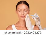 Small photo of Young caucasian woman getting hyaluronic acid filler for her nasolabial folds, getting rid of mimic wrinkles, standing over beige studio background, copy space