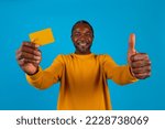 Small photo of Cheerful middle aged black man in yellow showing yellow bank credit card and thumb up, recommending unlimited shopping, contactless payment, isolated blue studio background