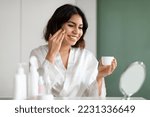 Skin Care Concept. Beautiful Female Using New Cosmetic At Home, Attractive Middle Eastern Young Woman In White Silk Robe Sitting In Front Of Mirror And Applying Moisturising Face Cream, Copy Space