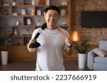 Small photo of Active sporty lifestyle. Excited japanese middle aged man holding trx resistance bands hang around his neck, standing in living room and posing to camera