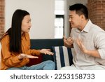 Angry asian spouses having quarrel, sitting on sofa and arguing, looking at each other, side view. Domestic violence and abuse. Couple struggling from marital crisis concept