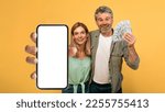 Small photo of Giveaway concept. Emotional middle aged couple holding a lot of dollar cash and showing smartphone with blank screen, posing over yellow studio background, mockup