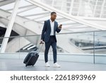 Portrait Of Handsome Black Businessman Walking With Suitcase In Airport And Using Smartphone, Young African American Man In Suit Browsing Internet On Cellphone While Going To Flight Gate, Copy Space