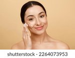 Small photo of Beautiful Young Indian Female Applying Moisturizing Cream On Face, Attractive Millennial Hindu Woman Nourishing Skin While Standing With Bare Shoulders Over Beige Studio Background, Copy