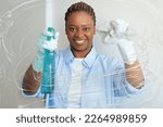 Small photo of Cheerful smiling funny young african american lady in comfortable outwear and rubber gloves cleaning windows at home, holding spray and cloth, black woman enjoyin chores