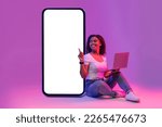 Online Offer. Happy black woman with laptop pointing at big blank smartphone with white screen while sitting in neon light over purple studio background, smiling lady recommending new app, mockup