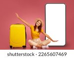 Small photo of Excited happy pretty young woman in hat and comfy casual outwear sitting on floor by yellow suitcase and big cell phone with white screen, imitating airplane, pink studio background, mockup