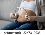 Unrecognizable Pregnant Woman Holding Wireless Headphones Near Belly, Expectant Lady Using Earphones For Playing Music To Baby In Womb While Sitting On Couch At Home, Closeup Shot With Free Space
