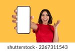 Small photo of Emotional pretty young arab woman in red t-shirt showing brand new cell phone with white empty screen and gesturing over yellow studio background, online offer, mockup, panorama