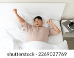 Happy asian middle aged man enjoying good morning, slying in comfy bed in white cozy bedroom, stretching his body after waking up, feeling excited and well-rested, top view, copy space