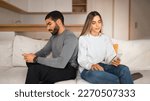 Small photo of Offended sad millennial arabic guy ignores european lady, watch video on phone in room interior, panorama. Chat and blog app, gadget addiction, treason online and problems in relations, bored at home