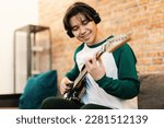 Cheerful Asian Student Guy Playing Electric Guitar Wearing Earphones Sitting On Sofa In Modern Living Room At Home. Boy Learning New Chords On Weekend. Hobby And Musical Gadgets. Selective Focus