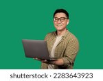 Positive asian man holding laptop computer and smiling at camera, wearing earpods and eyeglasses, posing over green background. Entrepreneurship career and internet technology concept