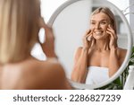 Small photo of Anti-Aging Cosmetics. Beautiful Middle Aged Woman Applying Eye Cream While Standing Near Mirror At Home, Attractive Mature Lady Touching Her Face, Enjoying Daily Beauty Routine, Selective Focus