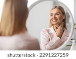 Beautiful Middle Aged Woman Looking At Mirror And Touching Her Soft Skin On Neck, Attractive Mature Lady Smiling To Reflection, Enjoying Anti-Aging Skincare Routine At Home, Selective Focus
