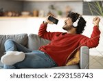 Domestic Fun. Happy Indian Man Listening Music In Wireless Headphones And Singing With Smartphone At Home, Cheerful Young Eastern Guy Relaxing On Couch In Living Room, Enjoying Favorite Playlist