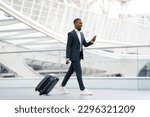 Handsome black businessman walking with suitcase at airport and using smartphone, young african american male wearing suit browsing internet on cellphone or messaging while going to flight boarding